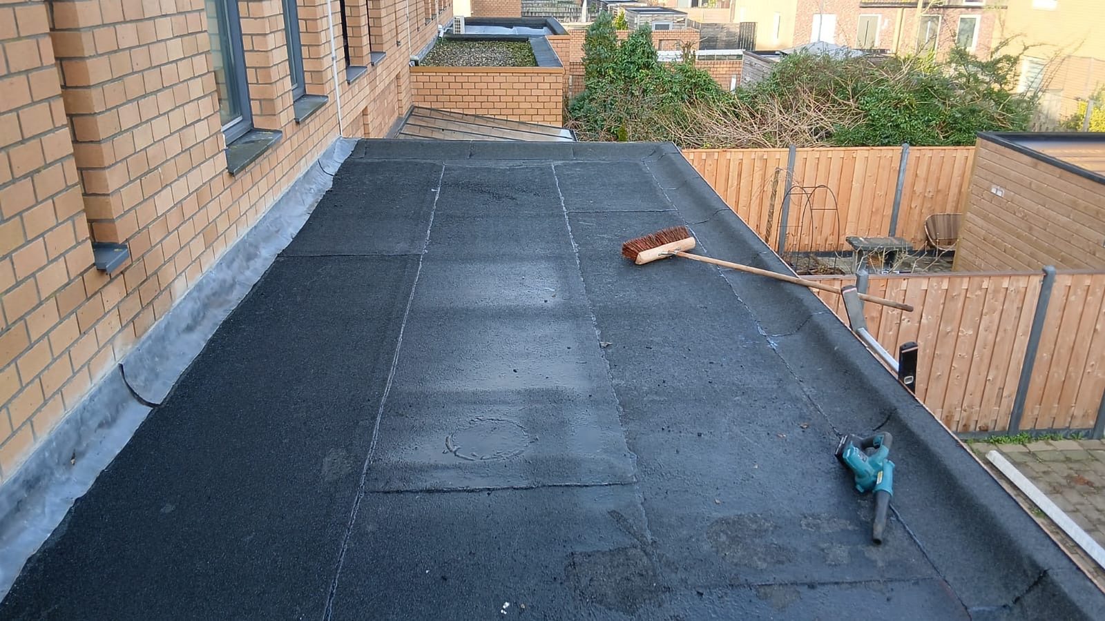Afgewerkt plat bitumen dak langs een gemetselde gevel, met bezem en gereedschap op het dakvlak