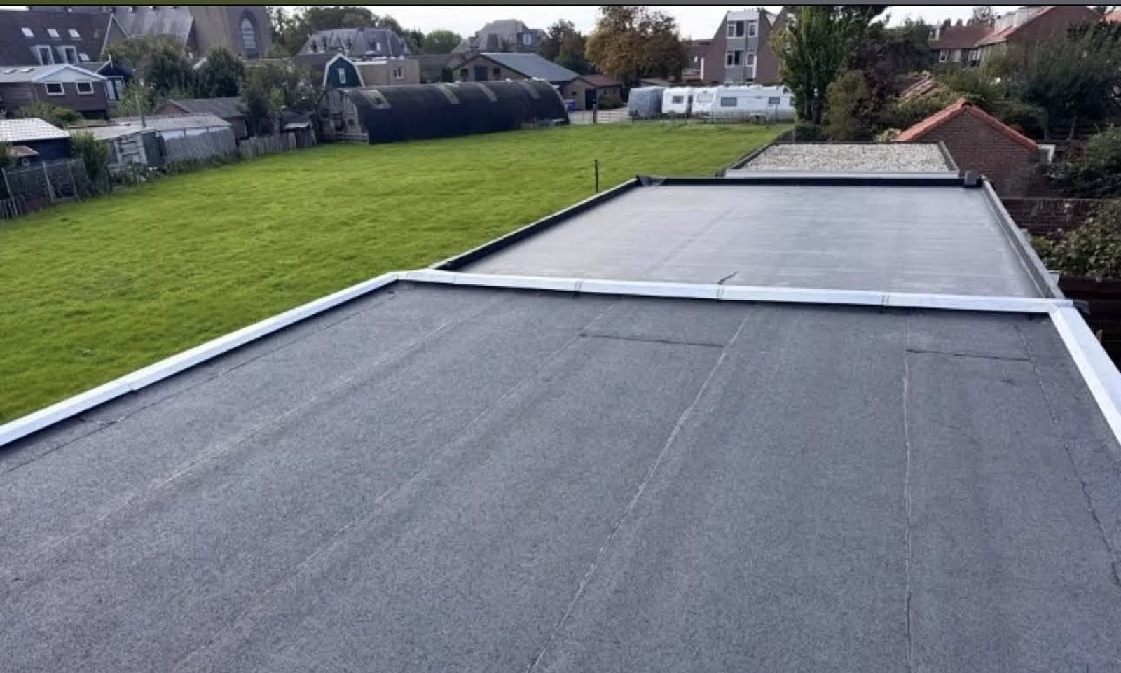Groot plat bitumen dakvlak met zinken deklijsten langs de randen en uitzicht op een grasveld